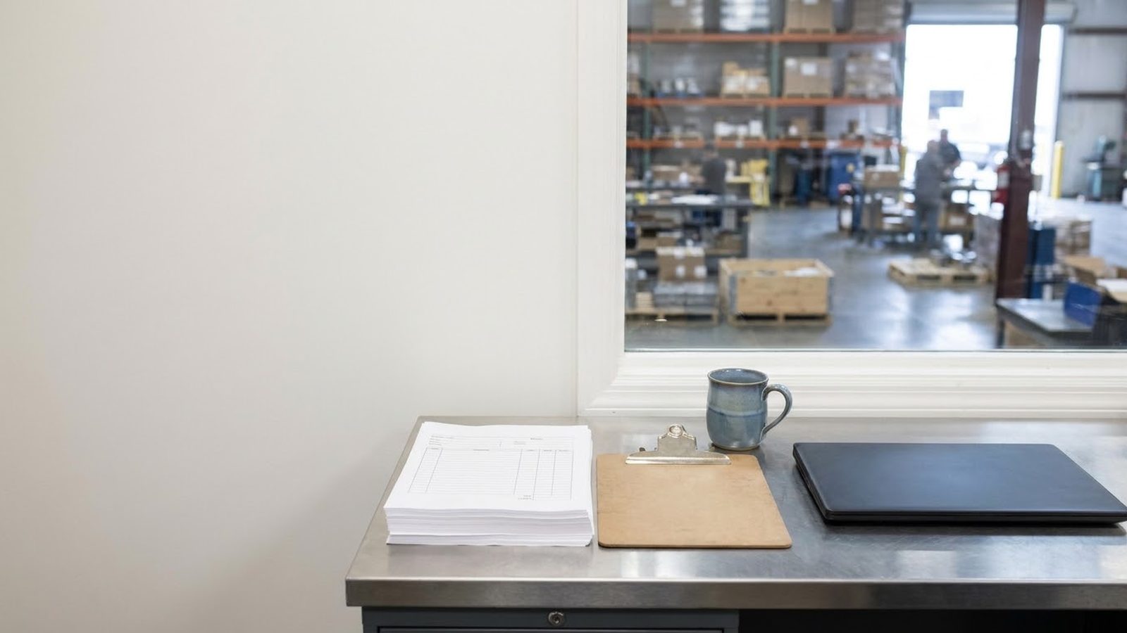 AI automation agency work for a manufacturer: desk with packing slips, a clipboard and a laptop overlooking the warehouse floor
