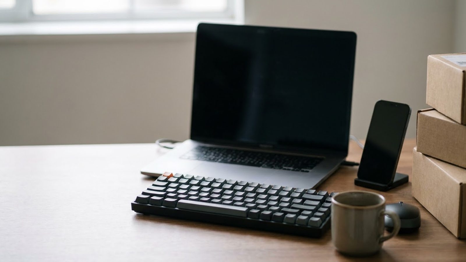Shopify app development company desk: open laptop, keyboard and a phone on a stand beside stacked shipping boxes